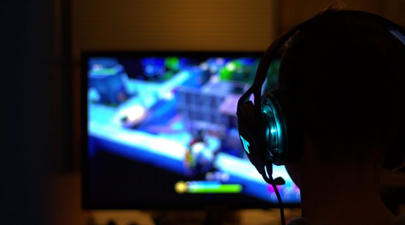 mężczyzna grający na komputerze, na ekranie jakaś gra, mężczyzna ma słuchawki, jest ciemno w pomieszczeniu, ekran rozświetla pomieszczenie