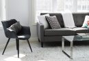 Skromne mieszkanie - krzesło, sofa i stolik. Biały dywan i białe ściany, szare meble. Za sofą znajduje się okno, widok na jakiś budynek